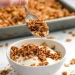 Hand sprinkling freshly made granola over a bowl of yogurt, with a baking tray of granola in the background